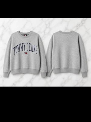 Grå sweatshirt från Tommy Jeans - Klassisk grå sweatshirt från Tommy Jeans med rund halsringning och tryckt logga i marinblått framtill. Tröjan har ribbade muddar vid ärmslut och nederkant samt en liten logodetalj på ärmen. Perfekt för en avslappnad och stilren look.