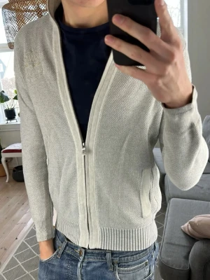 Ljusgrå Gant ziptröja med brodyr - Ljusgrå stickad ziptröja från Gant med broderade detaljer på axel och ärm. Tröjan har ribbade muddar, ståkrage och två fickor framtill. Perfekt för lager-på-lager och enkel att matcha med jeans eller chinos.