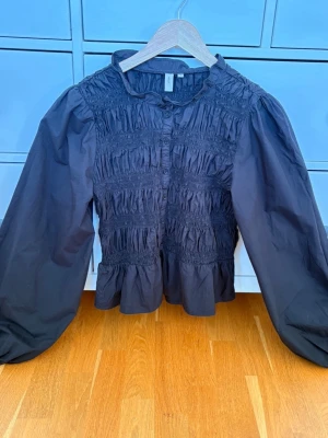 Svart smockad blus från Nelly - Trendig svart blus från Nelly omed smockade detaljer över hela bröstet och magen. Blusen har knappar framtill, volang nertill och långa puffiga ärmar. Perfekt för dig som gillar en cool och stilren look.