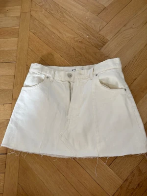 Vit jeanskjol från Pull&Bear - Snygg vit kort jeanskjol från Pull&Bear med rå kant nertill, klassiska fickor och bälteshällor. Kjolen har knapp och dragkedja framtill och är perfekt för en trendig look.