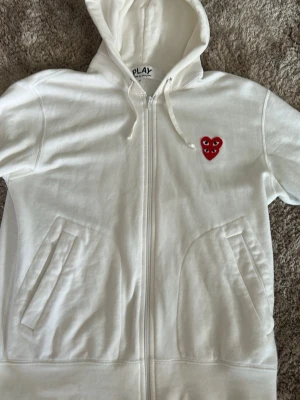 Comme des Garçons Play (cdg) hoodie - Vit hoodie med dragkedja från Comme des Garçons Play. Har huva med dragkedja, två fickor fram och det ikoniska röda hjärtat med ögon broderat på bröstet. Perfekt för en clean och stilren look.