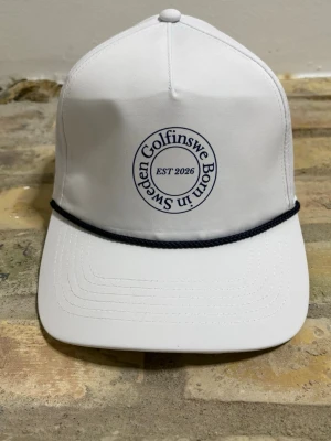 Vit golf rope keps Golfinswe  - Vit keps från Golfinswe med broderad text 'Born in Sweden' och 'EST 2026' framtill. Klassisk form med böjd skärm och svart snöre ovanför skärmen. Perfekt accessoar för en sportig och stilren look.