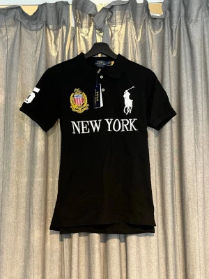 Svart pikétröja Polo Ralph Lauren - Svart pikétröja från Polo Ralph Lauren med vit broderad logga, texten NEW YORK framtill och nummer 5 på ärm och rygg. Klassisk krage och korta ärmar. Broderad patch med amerikansk flagga på bröstet. Perfekt för en sportig och stilren look.