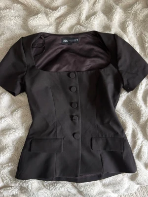Svart topp med knappar från Zara - Stilren svart topp från Zara med korta ärmar, markerad midja och stora klädda knappar framtill. Toppen har två dekorativa fickor och en fyrkantig urringning som ger en modern look. Perfekt att styla med jeans eller kjol.