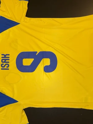 Sverige fotbollströja Isak 9 Adidas - Gul och blå officiell Sverige fotbollströja från Adidas med Isak och nummer 9 på ryggen. Tröjan har korta ärmar, broderad landslagssköld och Adidas-logga på bröstet. Perfekt för dig som vill stötta landslaget på match eller träning.
