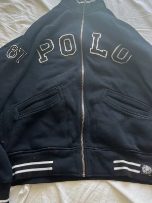 Svart helzip från Polo Ralph Lauren - Svart helzip från Polo Ralph Lauren. Stora vita bokstäver 'POLO' och siffran 67 på ena armen, två fickor och vita ränder på muddar och krage. Klassisk streetstil med collegevibbar och broderad logga vid nederkanten. Passar någon som är runt 180