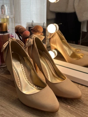 Beige pumps med rosett - Snygga beige pumps med spetsig tå och hög klack. Skorna har en dekorativ rosett baktill och insida i guldigt material. Perfekta för att lyfta din outfit med en feminin touch.