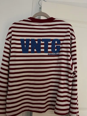Randig långärmad topp VNTG - Säljer en vit och vinröd randig långärmad topp med blå VNTG-tryck på ryggen. Toppen har en relaxed passform och breda ärmar, samt rund halsringning med vinröd kant. Perfekt för en chill och trendig look.