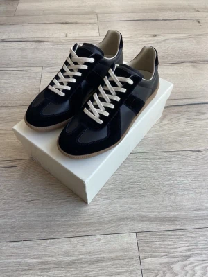 Maison margiela gats storlek 45 - Stilrena svarta sneakers med vita snören och beige sula. Skorna har en mix av mocka och slät läderlook, vilket ger en snygg kontrast. Låg modell som passar perfekt till jeans eller chinos. Klassisk design med sportig känsla.