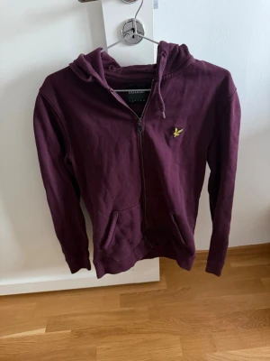 Vinröd hoodie från Lyle & Scott - Snygg vinröd hoodie med dragkedja från Lyle & Scott. Klassisk modell med huva, känguruficka och gul logga på bröstet. Mjuk bomullsblandning som är skön att ha på sig. Perfekt för en avslappnad och stilren look.