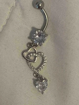 Navelpiercing med hjärtan och stenar - Snygg navelpiercing i silverfärgad metall med två gnistrande stenar och två hjärtan, varav ett är dekorerat med små glittrande stenar. Perfekt accessoar för att ge din look lite extra bling.