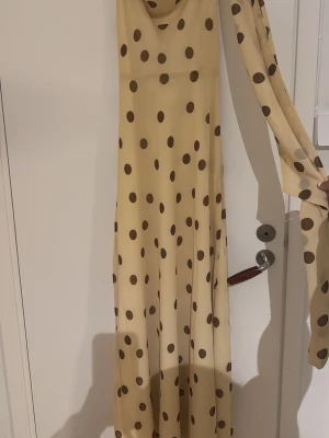 Beige långklänning med bruna prickar - Säljer en ljus gul långklänning med bruna prickar och transparenta långa ärmar. Klänningen har en mjuk, flytande passform och är tillverkad i ett tunt, lätt material som ger ett elegant intryck. Perfekt för dig som vill sticka ut med ett klassiskt prickigt mönster.