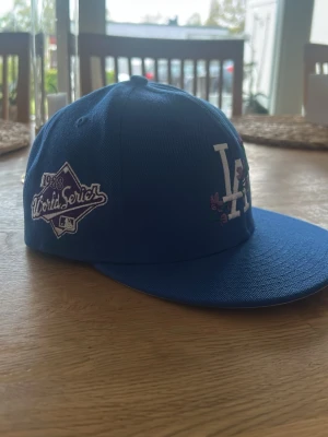 Blå LA Dodgers keps 59FIFTY - Snygg blå LA Dodgers keps från New Era 59FIFTY-serien med broderad 1988 World Series-logga på sidan och LA-logga framtill med lila blommor. Klassisk flat peak och broderade detaljer. Perfekt för dig som gillar sportig streetstyle.