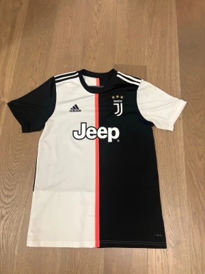 Juventus matchtröja Adidas M - Juventus officiella matchtröja från Adidas i svart och vitt med röd rand i mitten. Tröjan har klubbmärke, tre stjärnor, Jeep-logga och Adidas-logga på bröstet. Tillverkad i Climalite-material som andas, perfekt för fotbollsträning eller match.