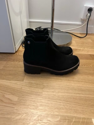 Svarta Chelsea boots i mocka - Säljer ett par svarta Chelsea boots i mocka med grov sula och elastiska paneler på sidorna. Klassisk siluett med dragflik bak och snygg kontrastsöm runt sulan. Mycket skick