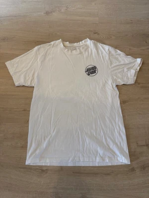 Vit Santa Cruz t-shirt med tryck - Cool vit t-shirt från Santa Cruz med svart logotyp på bröstet och ett stort grafiskt tryck med en hand och döskalle på ryggen. Klassisk passform och rund hals. Perfekt för dig som gillar streetwear och skatekultur.