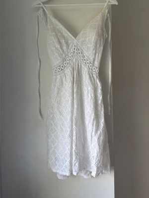 Vit spetsklänning med öppen rygg - Söt vit kortklänning i spets med v-ringning och smala axelband. Klänningen har ett fint broderat mönster, markerad midja och öppen rygg. Perfekt för sommarens alla tillfällen. Knytband i sidorna för extra detalj.
