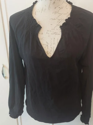 Svart blus med smockad krage - Svart blus med lång ärm och smockad, rynkad krage samt ärmslut. Djup v-ringning framtill och mjukt, lätt tyg som ger en avslappnad men stilren look. 