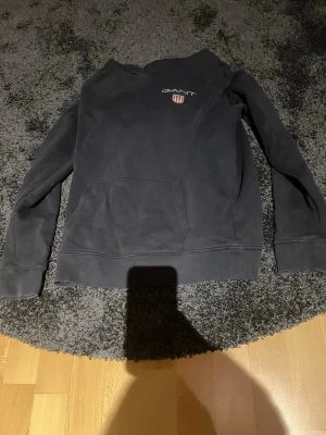 Svart hoodie från GANT med logga - Svart hoodie från GANT med broderad logga och sköld på bröstet. Klassisk känguruficka framtill och mjukt material som känns skönt mot huden. Perfekt för en avslappnad och stilren look.