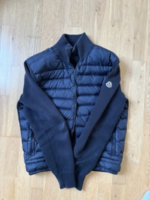 Mörkblå cardiganjacka från Moncler - Snygg mörkblå cardiganjacka från Moncler med quiltad front och stickade ärmar. Jackan har dragkedja, hög krage och Moncler-logga på ärmen. Perfekt för lager-på-lager och streetstyle. Materialmix av dun och stickat ger både värme och stil.
