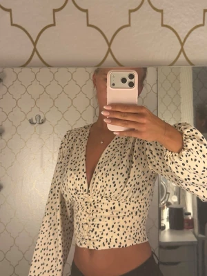 Prickig croppad blus med v-ringning - Snygg croppad blus i ljus färg med svarta prickar och v-ringning, från Gina tricot i storlek 34. Blusen har långa puffiga ärmar och markerad midja som ger en trendig siluett. Perfekt för dig som vill sticka ut med en cool och lekfull stil.