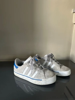 Grå Adidas Campus sneakers med blå detalj - Snygga grå sneakers från Adidas med klassiska vita ränder på sidorna och en blå detalj vid hälen. Skorna har en låg profil, grå mocka och vita snören. Perfekta för en avslappnad streetstyle-look.