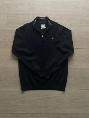 Svart half zip stickad tröja Gant - Svart stickad tröja från Gant med halv dragkedja och ribbad krage med vita ränder. Liten broderad logga på bröstet. Långärmad modell i mjukt material, perfekt för lager-på-lager-stil. Tröjan är sjukt skön och väl omhändertagen!