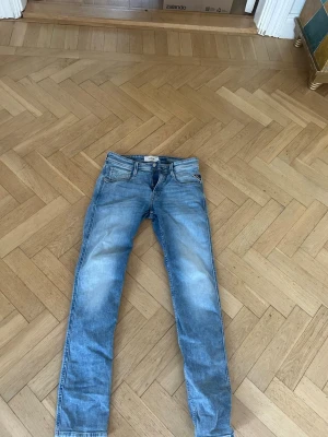 Ljusblå slim fit jeans från Replay - Snygga ljusblå slim fit jeans från Replay med klassisk femficksdesign och dragkedja fram. Jeansen har en tvättad look med slitningar och en brun läderpatch med logga bak i midjan. Perfekta för en avslappnad och trendig stil.