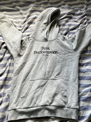 Grå Peak Performance Hoodie  - grå hoodie från Peak Performance med svart broderad logga på bröstet. Perfekt för en avslappnad och sportig stil. Mjuk bomullsblandning som är skön att ha på sig.