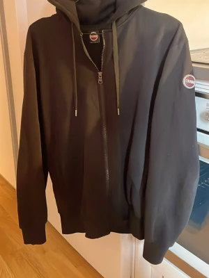 Svart hoodie med dragkedja från Colmar - Snygg svart hoodie från Colmar med dragkedja framtill och justerbar huva. Märkeslogga på bröstet och ärmen. Perfekt för en avslappnad och stilren look. Mjukt material och ribbade muddar vid ärmslut och nederkant. Passar M