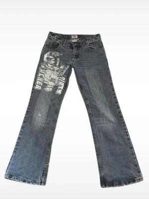 Blå bootcut jeans med tryck - Säljer ett par blå bootcut jeans med stort vitt grafiskt tryck på ena benet och bakfickan. Jeansen har klassisk femficksmodell, låg midja och slitna detaljer. Perfekta för dig som vill sticka ut med en unik streetwear-look. de är omsydda lite lägre.