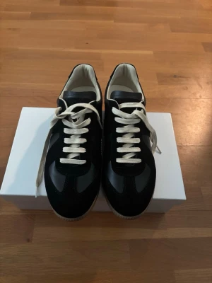 Maison Margiela Gats Black - Stilrena svarta sneakers med beige sula. Ovandelen är i en mix av mocka och läder, vilket ger en snygg kontrast och premiumkänsla. Insidan är ljus och skorna har en klassisk låg profil som passar till många outfits.