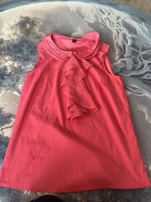 Rosa ärmlös blus med volang - Säljer en söt rosa ärmlös blus med dekorativ volang framtill och rund krage med knappdetalj. Perfekt för dig som gillar en feminin och stilren look. Blusen har en lätt och luftig känsla, vilket gör den skön att bära under varma dagar.