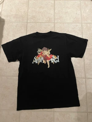 Amiri Tshirt - Svart t-shirt från Amiri med stort tryck av en ängel i röd klänning och guldiga detaljer över bröstet. T-shirten har klassisk passform och rund halsringning. Perfekt statement-plagg för dig som gillar unika prints.