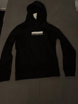 Svart hoodie från Calvin Klein Jeans - Svart hoodie från Calvin Klein Jeans med vit logga på bröstet. Klassisk modell med huva och långa ärmar. Tillverkad i mjukt material som är skönt att bära. Perfekt för en avslappnad och stilren look.