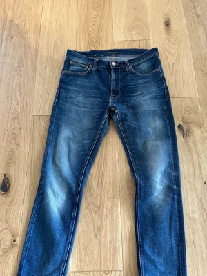 Blå slim fit jeans från Nudie Jeans - Säljer ett par blå slim fit jeans från Nudie Jeans med riktigt snygga slitningar/tvätt och klassisk femficksmodell. Jeansen har orangea sömmar och är tillverkade i 99% bomull 1% elastane. Perfekta för en avslappnad och trendig look. 