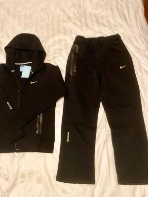 Svart Nike NOCTA loungewear set - Snyggt svart loungewear set från Nike NOCTA med gul broderad Swoosh och NOCTA-logga på både hoodie och byxor. Hoodien har dragkedja, huva och fickor med svarta detaljer. Byxorna har dragsko i midjan och raka ben. Perfekt för chill dagar hemma.