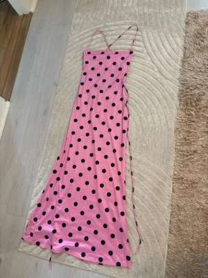 Rosa långklänning med svarta prickar - Supersöt långklänning i rosa med svarta prickar och tunna axelband. Klänningen har en figurnära passform och är tillverkad i ett mjukt, glansigt material som ger en lyxig känsla. Perfekt för dig som vill sticka ut med en lekfull och trendig look.