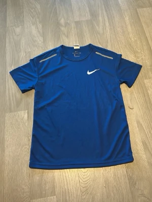 Blå Nike tränings-tshirt finns i mer färger kom dm   - Snygg blå tränings-tshirt från Nike med vit logga på bröstet och reflekterande detaljer på axlarna. Tillverkad i ett lätt och ventilerande funktionsmaterial som passar perfekt till träning och sportaktiviteter. Klassisk rund hals och korta ärmar.