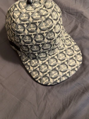 Mönstrad keps från Salvatore Ferragamo - Snygg keps från Salvatore Ferragamo med grått och vitt mönster över hela kepsen. Justerbar rem bak med metallspänne och svart läderdetalj med logga. Tillverkad i tygmaterial, perfekt för att lyfta din streetstyle.