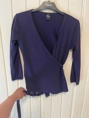 Lila omlottopp från Filippa K - Snygg lila omlottopp från Filippa K med trekvartsärm och knyt i midjan. Tillverkad i mjuk bomull med lite stretch för en bekväm passform. Perfekt att styla med jeans eller kjol för en trendig look.