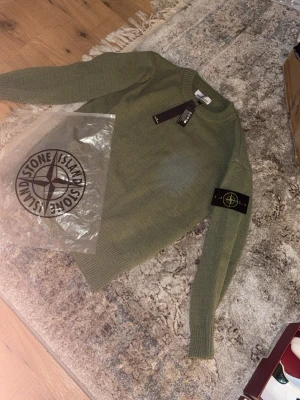 Grön stickad tröja från Stone Island - Säljer en snygg grön stickad tröja från Stone Island med klassisk patch på ärmen. Tröjan har rund halsringning och långa ärmar. Perfekt för dig som gillar streetwear och vill ha något stilrent och bekvämt.