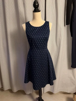 Mörkblå prickig ärmlös klänning - Snygg mörkblå klänning med vita prickar och ärmlös design. Klänningen har rund halsringning och en lätt utställd kjol som ger en feminin siluett. Perfekt för dig som gillar klassiska mönster och enkel stil.