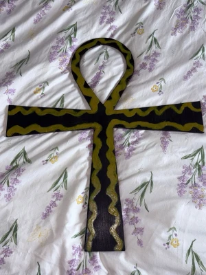 Ankh kors i trä med gul dekor - Säljer ett handgjort ankh kors i trä, målat i svart med gulgrön vågig dekor längs kanterna. Perfekt som accessoar till maskerad eller cosplay med egyptiskt tema. Korset har en klassisk ögla upptill och är robust i formen.