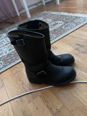 Svarta läderboots från Johnny Bulls - Klassiska svarta boots i läder från Johnny Bulls med grov sula och två spännen, ett vid skaftet och ett vid vristen. Bootsens höga skaft och robusta design ger en cool och tidlös look. Perfekta för dig som gillar en tuff stil.