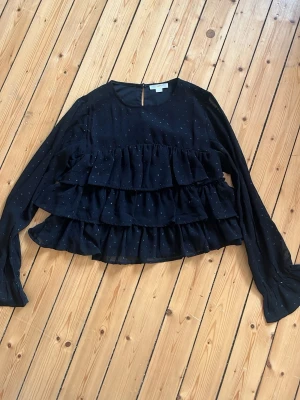 Svart volangblus från Gina Tricot - Svart blus från Gina Tricot med långa ärmar och flera volanglager framtill. Tunt, lätt material med små diskreta prickar som ger en extra detalj. Säljer pågrund av att den är för liten, jätte fin! Ordinarie pris 400☺️💗