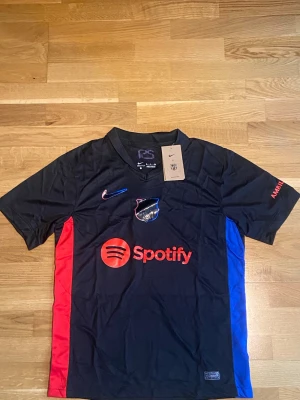Svart FC Barcelona Nike matchtröja - Snygg svart FC Barcelona matchtröja från Nike med röda och blå sidopaneler. Tröjan har korta ärmar, V-ringning och tryck med Spotify-logga på bröstet. Perfekt för fotbollsträning eller att bära som supporter. Materialet är lätt och andas.