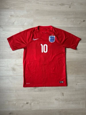 England Bortatröja 2014 Fotbollströja VM Rooney Storlek M  - Mycket fin fotbollströja i röd färg. Tröjan är i mycket bra skick och sparsamt använda. |  Äkta som allt annat jag säljer! ✅ |  Storlek: M | Skick: Mycket Bra Skick - Tyvärr har trycket börjat släppt litegrann på ryggen och lite syn på användning  (SE SISTA 2 BILDER) | Färg: Röd | Märke: Nike | Modell: Fotbollströja |   Hör gärna av dig vid frågor så svar