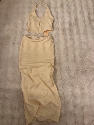 Ljusbeige lång kjol med topp - Trendig lång kjol i ljusbeige med omlott-design och lätt skrynkligt tyg. Kjolen har en rak passform och är perfekt för sommarens alla tillfällen. Matchande topp finns också om du vill köra på en hel look. Aldrig använd därav prislapp finns kvar. 