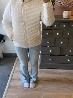 Vit pufferjacka med huva - Snygg vit pufferjacka med dragkedja framtill och två fickor på sidorna. Jackan har en mysig huva och quiltad design som håller värmen. Perfekt för kalla dagar och enkel att matcha med olika outfits.